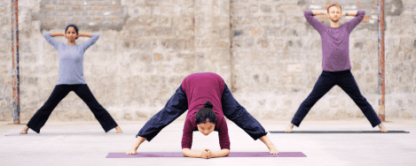 The image shows three individuals performing Angamardana yoga exercises in an outdoor setting against a stone wall. The person in the center is bent forward with legs spread wide apart, hands resting on the ground, and head down. The two individuals on either side are standing with legs spread wide and hands clasped behind their heads. They are all on yoga mats and appear to be engaged in a synchronized yoga session. This image is interesting and relevant as it depicts a group practicing Angamardana, a traditional Indian fitness system that involves a series of physical postures and movements aimed at strengthening the body and improving flexibility.