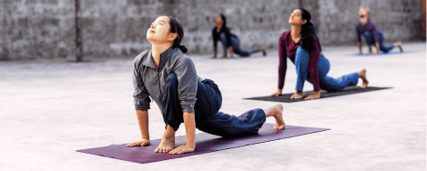 A group of individuals practicing yoga in an outdoor setting, each in a Ashvasanchalana pose, focusing on balance and flexibility. The participants, dressed in comfortable yoga attire, are on mats placed on a concrete floor with a simple, gray stone wall in the background. The image conveys focus, strength, and flexibility achieved by practicing Surya kriya & Surya shakti yogic practices.