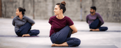 The image shows three individuals practicing yogasanas. They are seated on yoga mats in a cross-legged position with their torsos twisted to one side and their arms wrapped around their backs, performing a seated spinal twist pose. The background is a plain, outdoor setting with a concrete floor and a blurred wall. The individuals are dressed in comfortable, casual clothing suitable for yoga practice. This image is interesting and relevant as it depicts a common yoga pose that promotes flexibility and spinal health.