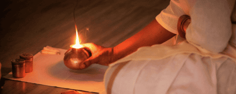 The image shows a close-up of a person performing a ritual, likely Bhuta Shuddhi, which is a traditional purification practice. The person is holding a small copper vessel with a flame coming out of it, indicating the use of fire in the ritual. The person is dressed in white clothing, and there are other small copper containers placed on a white cloth in front of them. The scene is dimly lit, emphasizing the glow of the flame and creating a serene atmosphere. This image is interesting and relevant as it captures a moment of spiritual practice, highlighting cultural and religious traditions.