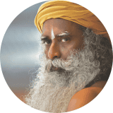 The image is a circular portrait of a person. The individual has long, curly white hair and is wearing a yellow turban, a traditional headgear. The background is a gradient of blue and gray colors, providing a calming backdrop. This image is of Sadhguru, a renowned spiritual leader, and yogi.