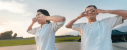The image shows two individuals standing outdoors, each performing the Shanmukhi mudra. The Shanmukhi mudra is a yogic hand gesture where the thumbs are used to close the ears, the index fingers close the eyes, the middle fingers close the nostrils, and the ring and little fingers press above and below the lips, respectively. Both individuals are wearing white shirts and are positioned against a backdrop of a clear sky and open field, with some trees and mountains visible in the distance. The image is interesting and relevant as it depicts a specific yogic practice aimed at sensory withdrawal and inner focus, which is significant in meditation and mindfulness practices.