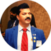 Student testimonial 1 The image shows an individual wearing a blue suit jacket, a white shirt, and a red tie with diagonal stripes. The individual has several pins and a name tag on the left side of the suit jacket. The background appears to be an indoor setting with another person partially visible behind the main subject. This image is used for a positive testimonial, likely highlighting the individual's professional appearance and achievements.